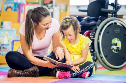 A young girl is sitting in front of her wheelchair. She is in an elementary school classroom with her helper. They are reading a story book together on the floor.